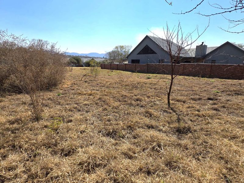 A view of a vacant residential plot featuring dry grass and a brick boundary wall, with neighboring houses visible in the background.