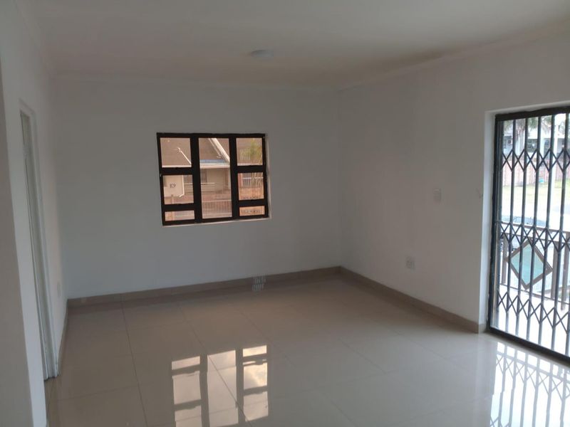 A bright, empty living area featuring neutral tiled flooring, a security gate, and an aluminium window frame, offering a clean blank canvas.