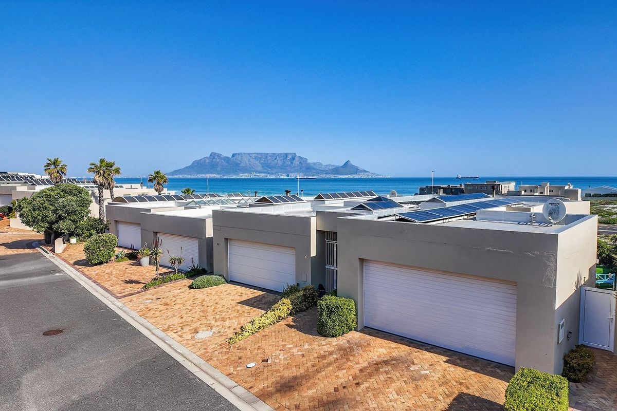 Aerial view of a residential complex featuring paved driveways, garage access, and rooftop solar panels with a scenic ocean and Table Mountain backdrop.