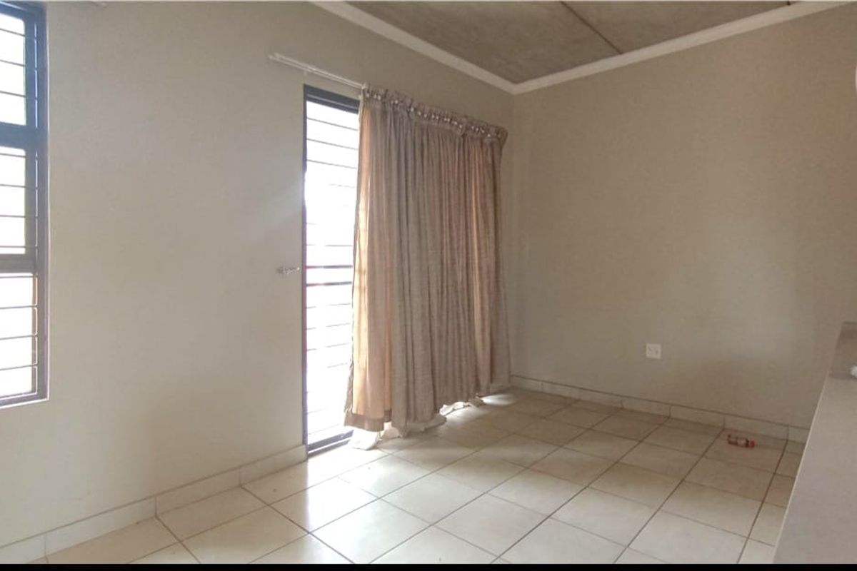 A simple, empty living area featuring tiled flooring, large windows with security bars, and neutral wall tones, offering a blank canvas for personalization.