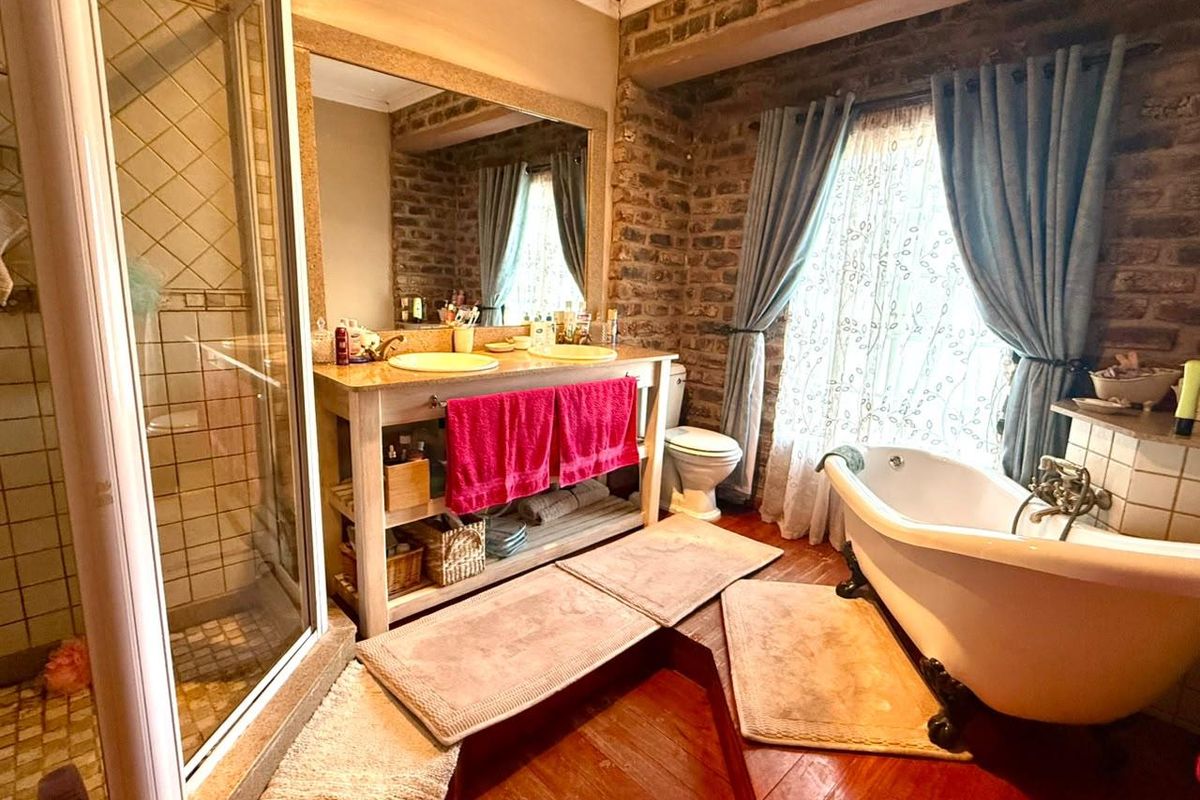 A rustic-style bathroom featuring an exposed brick wall, a freestanding clawfoot bathtub, a glass shower enclosure, and a double vanity with timber flooring.