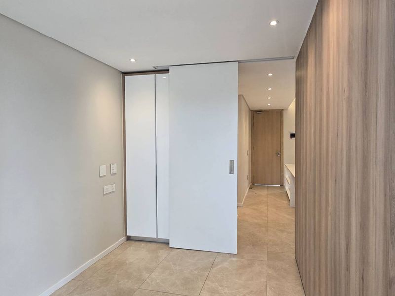 A modern, minimalist hallway featuring neutral tiled flooring, wood veneer wall paneling, and a sleek white sliding door leading to an adjacent room.