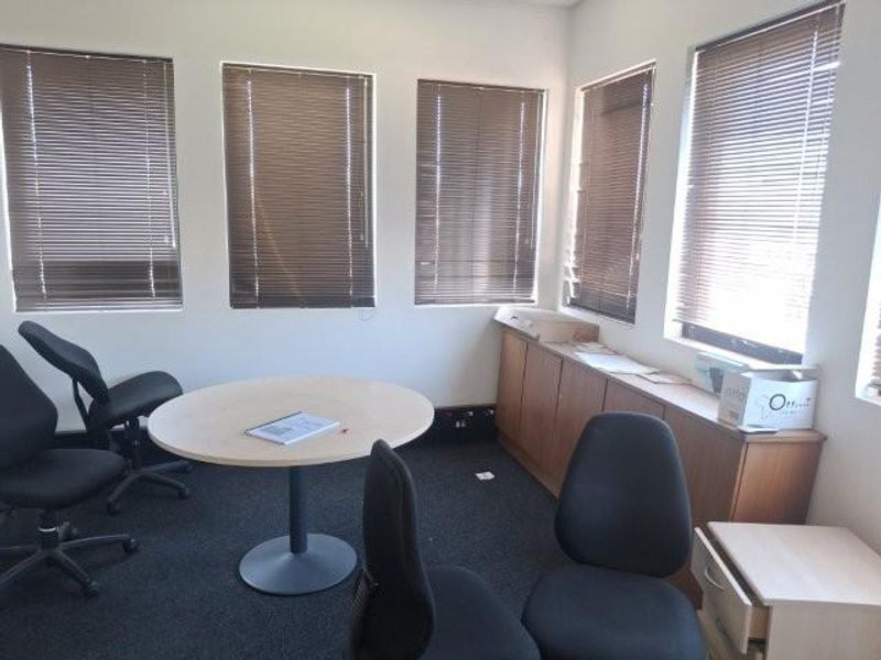 A functional office space featuring multiple windows with venetian blinds, built-in storage cabinetry, and dark carpeting.