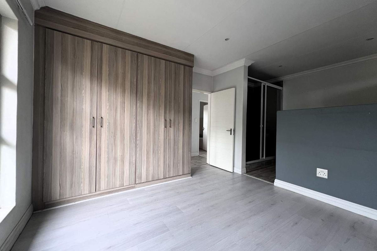 A bright bedroom featuring light wood-look laminate flooring, built-in floor-to-ceiling wooden cupboards, and a neutral grey accent wall.
