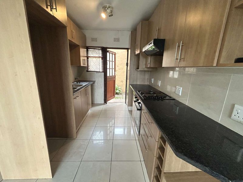 A functional galley-style kitchen featuring light wood cabinetry, dark granite countertops, built-in oven, gas hob, and tiled flooring with direct access to the exterior.