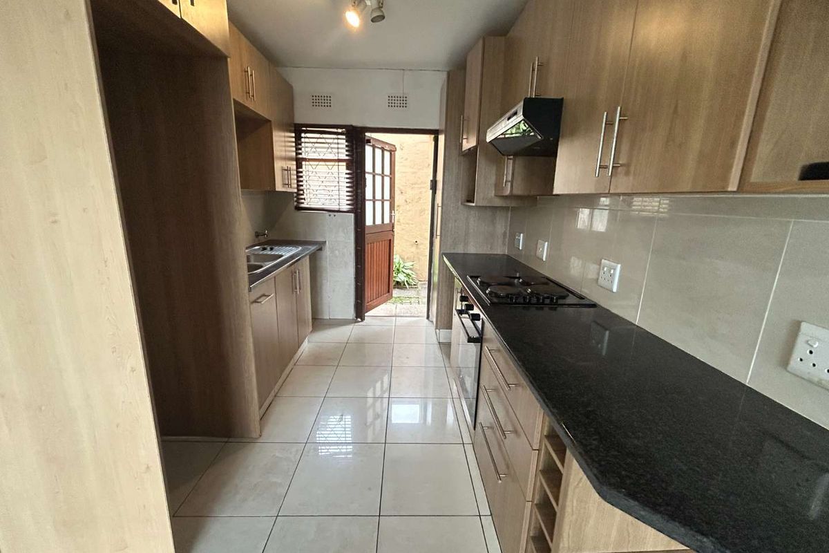 A functional galley-style kitchen featuring light wood cabinetry, dark granite countertops, built-in oven, gas hob, and tiled flooring with direct access to the exterior.