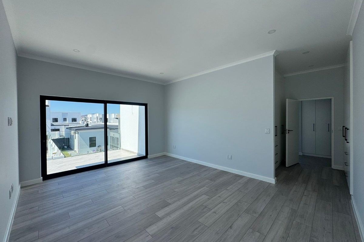 A bright, modern bedroom featuring light wood-look flooring, recessed ceiling lighting, and a large sliding glass door leading to an outdoor balcony.