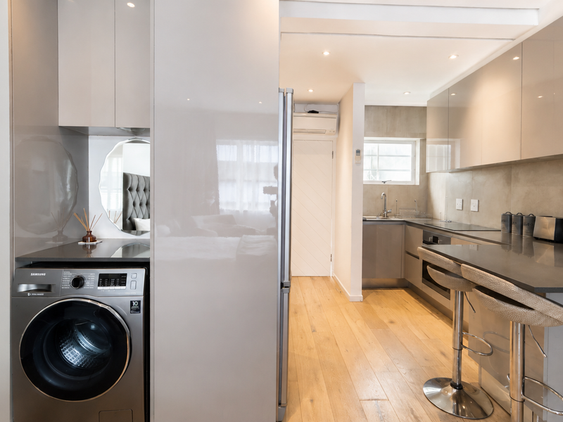 Modern kitchen featuring high-gloss cabinetry, stone countertops, integrated laundry appliance, and hardwood flooring, seamlessly flowing into the living area.