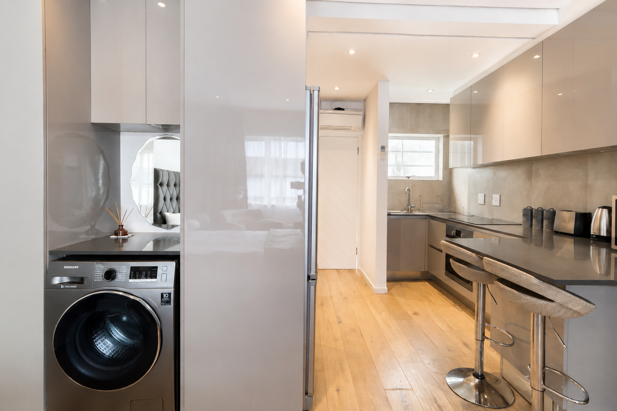 Modern kitchen featuring high-gloss cabinetry, stone countertops, integrated laundry appliance, and hardwood flooring, seamlessly flowing into the living area.