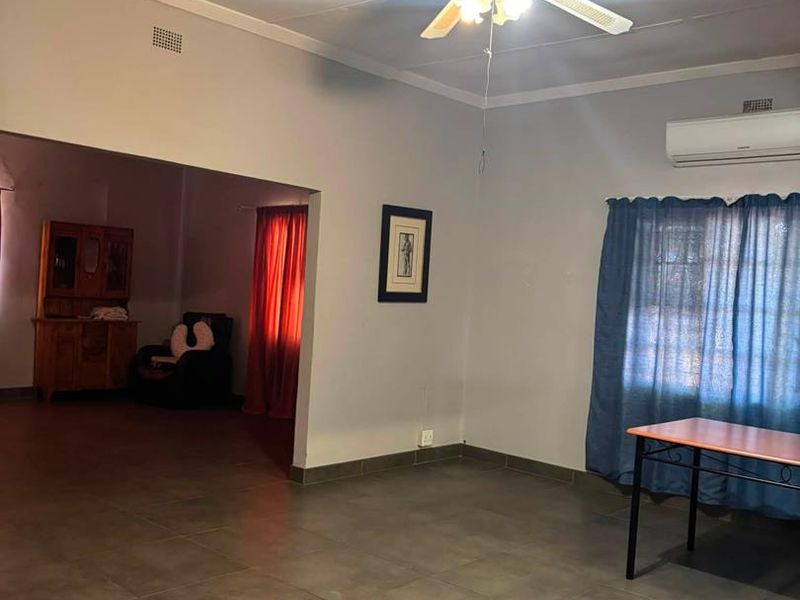 Spacious living area featuring tiled flooring, a ceiling fan, and a wall-mounted air conditioning unit, with an open-plan flow to an adjacent room.