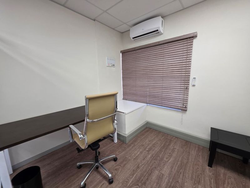 A functional office space featuring laminate flooring, a suspended ceiling, a wall-mounted air conditioning unit, and venetian blinds for light control.