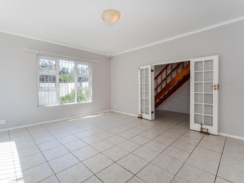 A bright, empty living room featuring neutral tiled flooring, large windows with security bars, and classic French doors leading to a staircase.