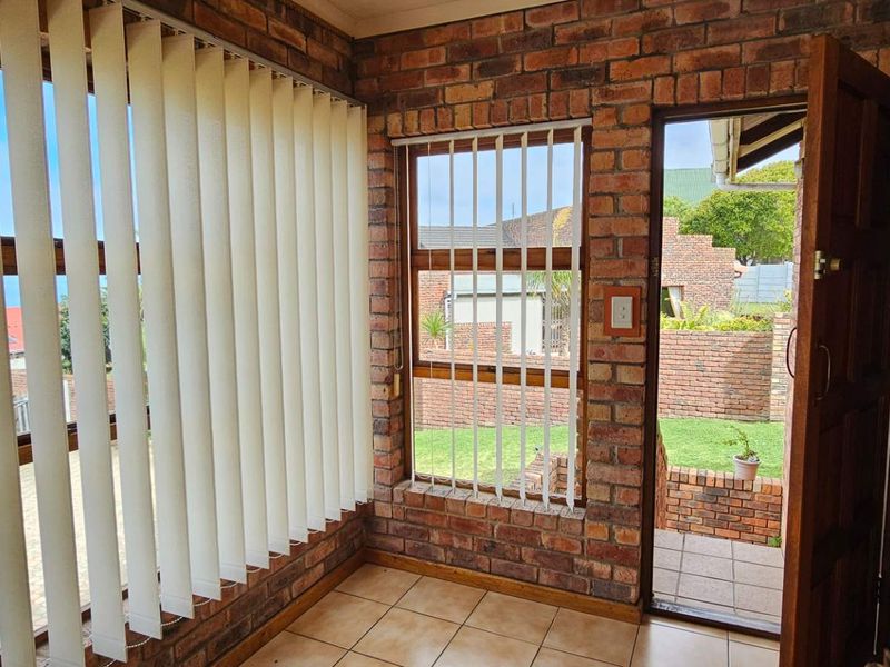 A sunroom or enclosed patio featuring exposed brick walls, tiled flooring, and large windows with vertical blinds, offering direct access to the garden area.
