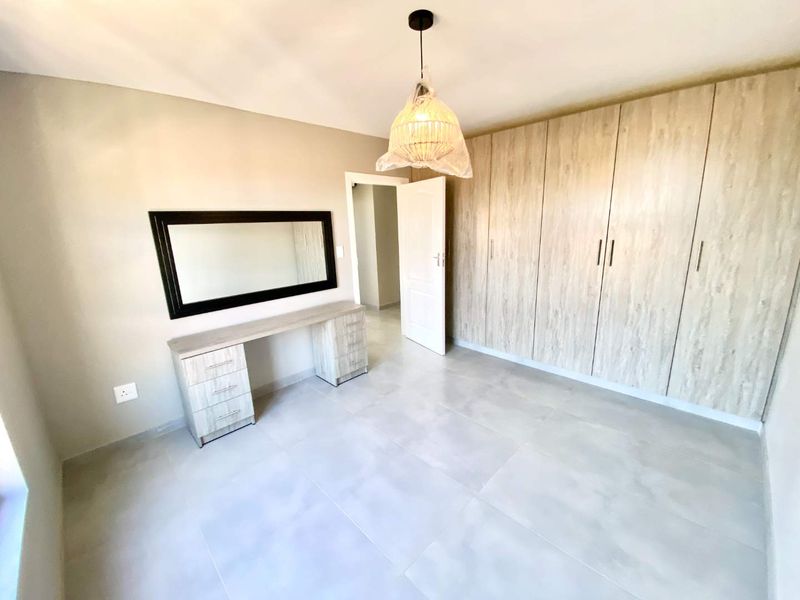 A bright, modern bedroom featuring neutral tiled flooring, extensive built-in wooden cupboards, and a matching built-in vanity unit.