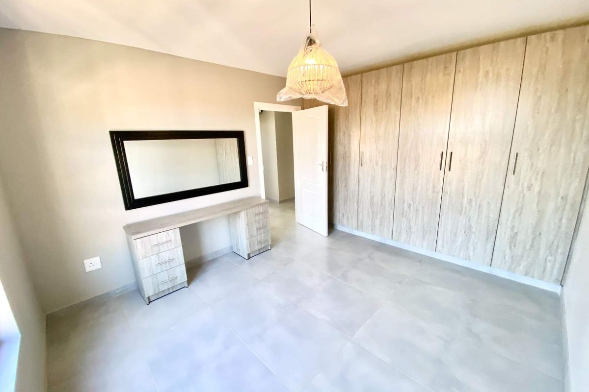 A bright, modern bedroom featuring neutral tiled flooring, extensive built-in wooden cupboards, and a matching built-in vanity unit.