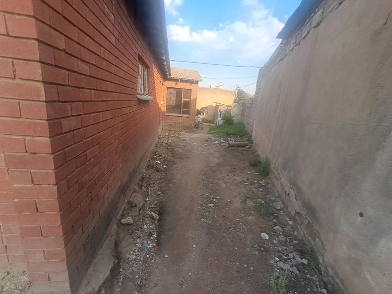 A narrow side passage between a brick house and a boundary wall, featuring unpaved ground and visible wear on the exterior surfaces.