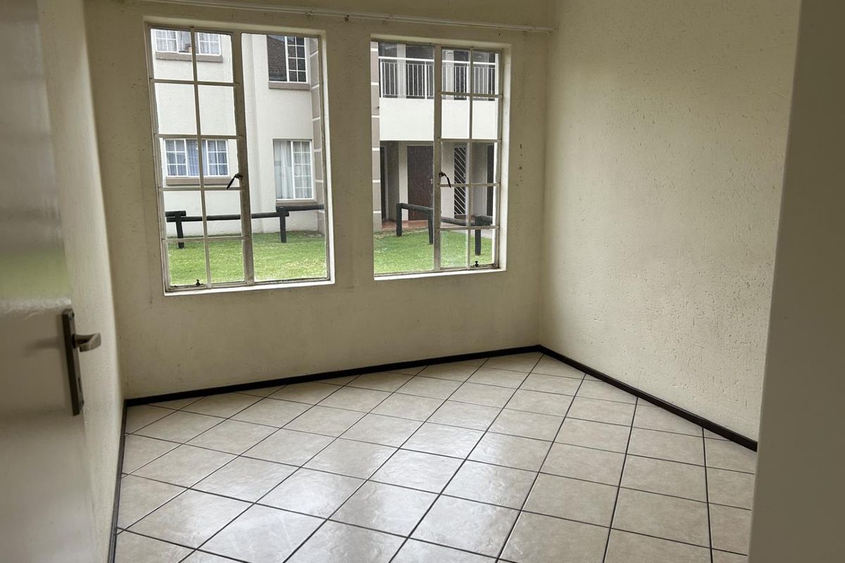 An empty bedroom featuring neutral tiled flooring and large steel-framed windows overlooking the complex grounds.