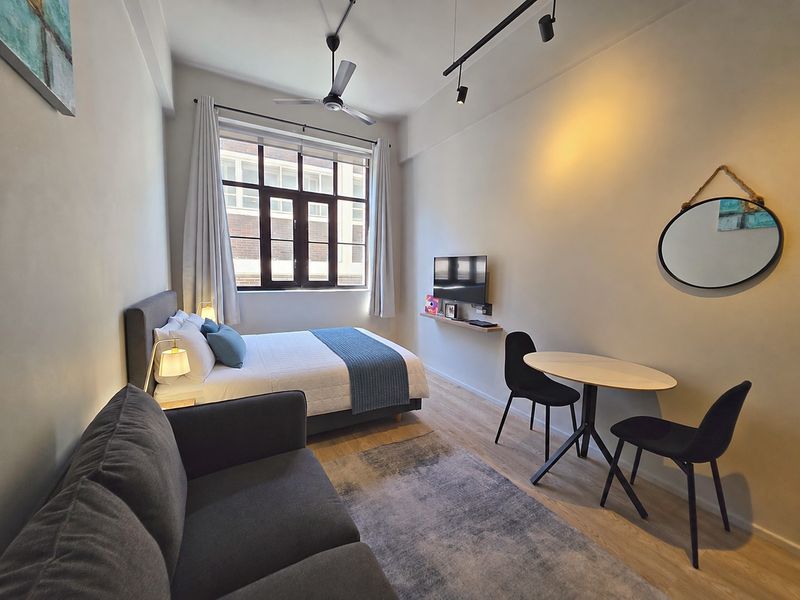 A well-maintained open-plan studio apartment featuring modern track lighting, a ceiling fan, and large windows that provide ample natural light.