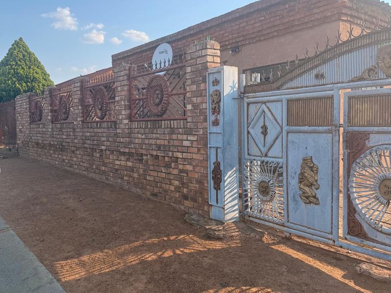 Front exterior view featuring a brick boundary wall with decorative metal fencing and a large, weathered metal entrance gate.