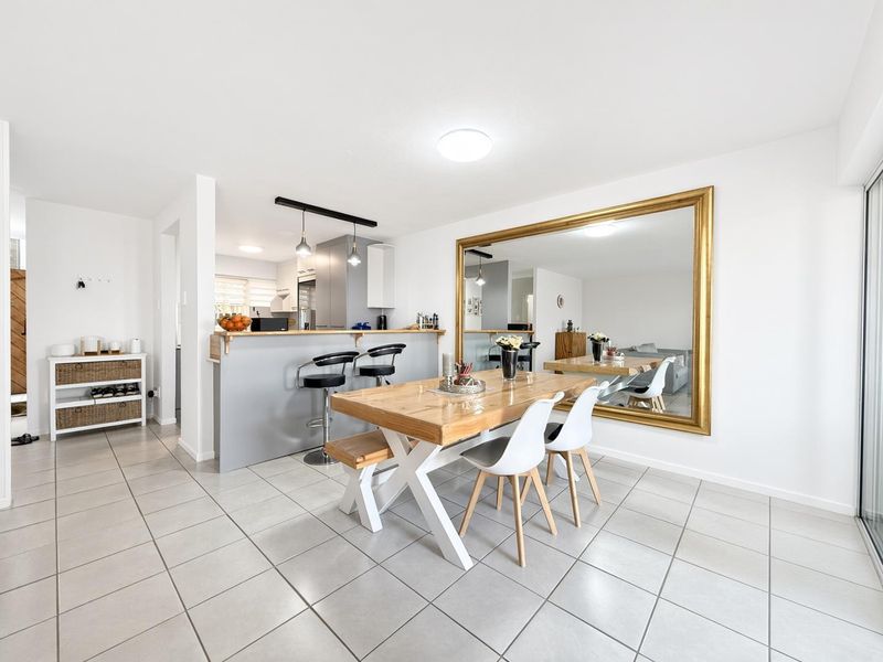 Bright open-plan dining and kitchen area featuring neutral ceramic floor tiles, a breakfast bar with pendant lighting, and a spacious layout.