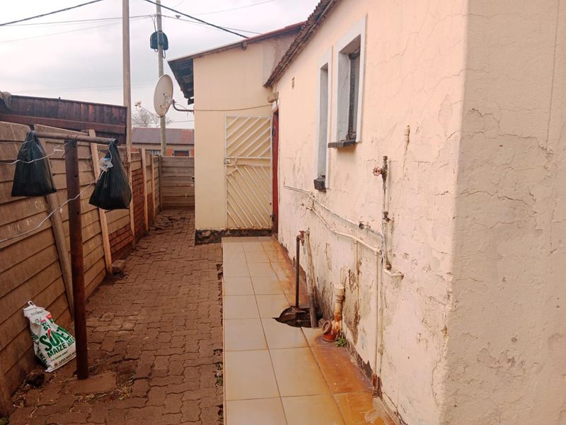 Rear exterior view of the property showing a paved walkway, security gate, and visible wear to the exterior wall plaster and flooring.