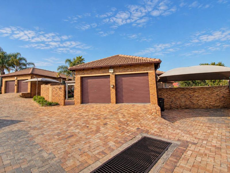 A well-maintained townhouse exterior featuring a double garage with roll-up doors, a paved driveway, and an additional shade cloth carport.
