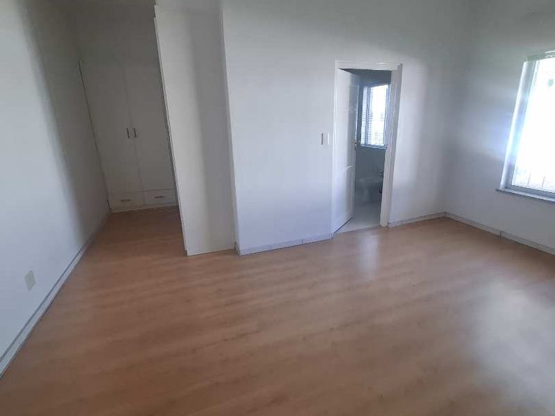 A spacious, empty bedroom featuring light wood-look laminate flooring, built-in cupboards, and recessed ceiling lighting, with an adjoining doorway.