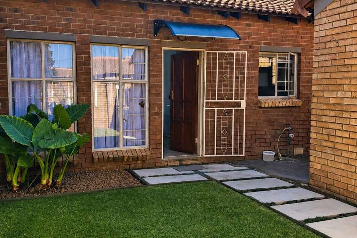 A rear exterior view of a brick townhouse featuring a small garden, paved walkway, and a security gate at the entrance.