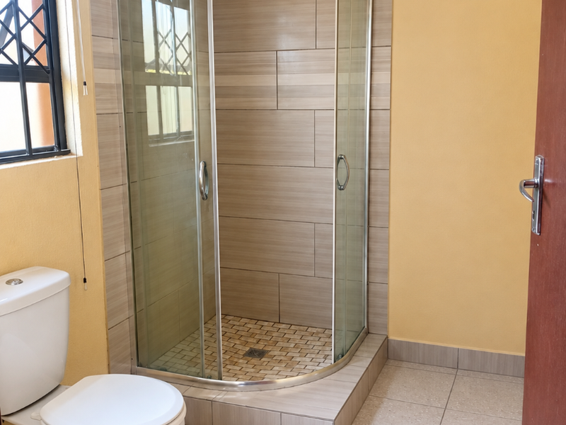 A clean, functional bathroom featuring a curved glass shower enclosure, neutral wall and floor tiling, and a standard toilet.