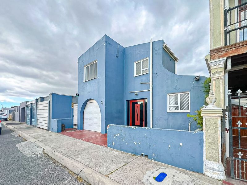 Front elevation of a blue-painted house featuring a single garage, red entrance door, and a low boundary wall with visible wear.