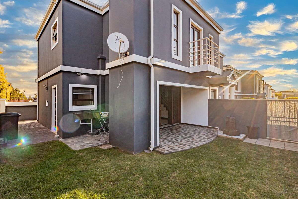 Rear view of a modern two-story home featuring a paved patio, small garden, built-in braai area, and a balcony with metal railing.