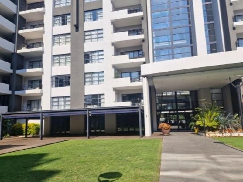 Modern apartment building exterior featuring a paved entrance walkway, landscaped lawn, covered patio area, and multi-level balconies with large glass windows.