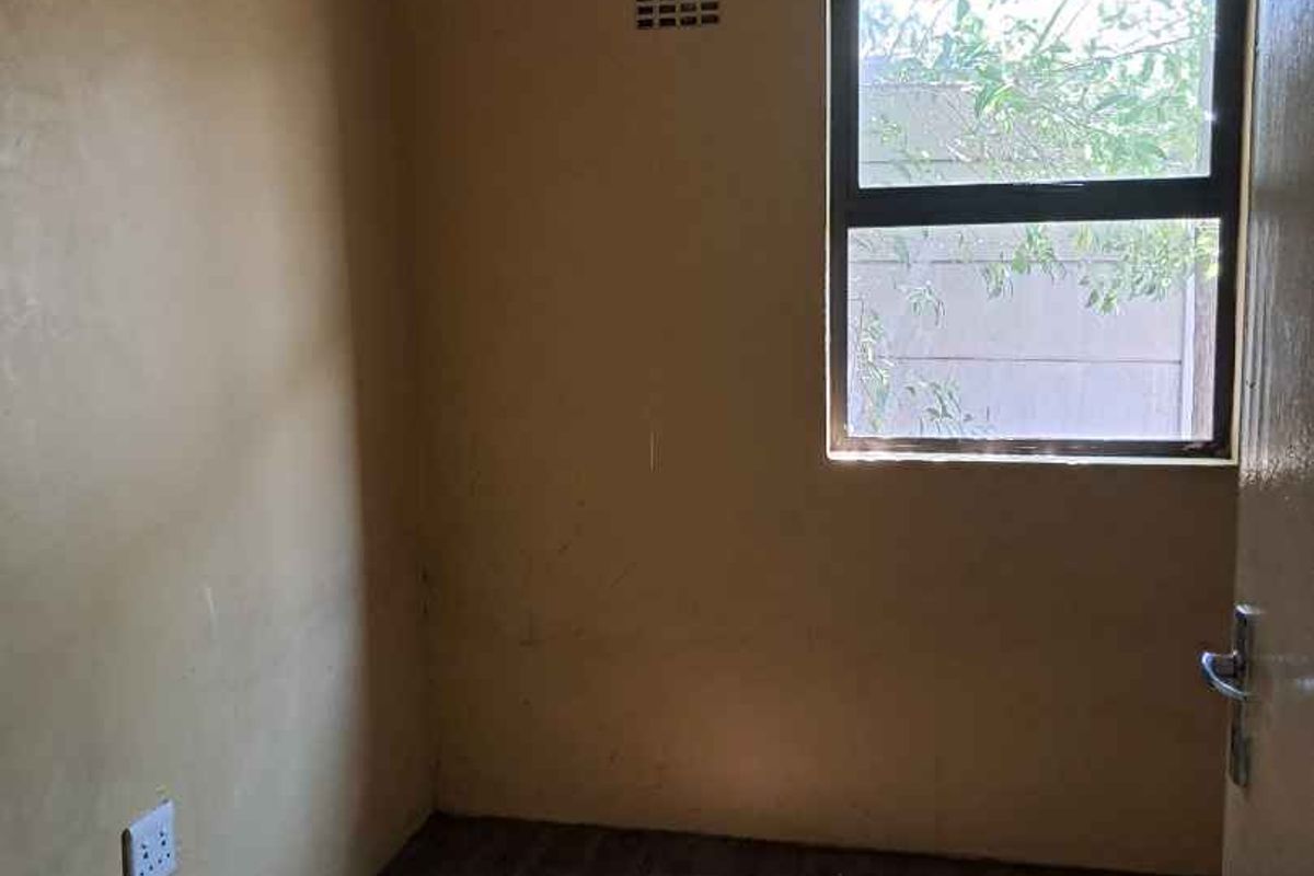 A small, empty bedroom featuring dark wood-look flooring, a single window with security bars, and plain painted walls.