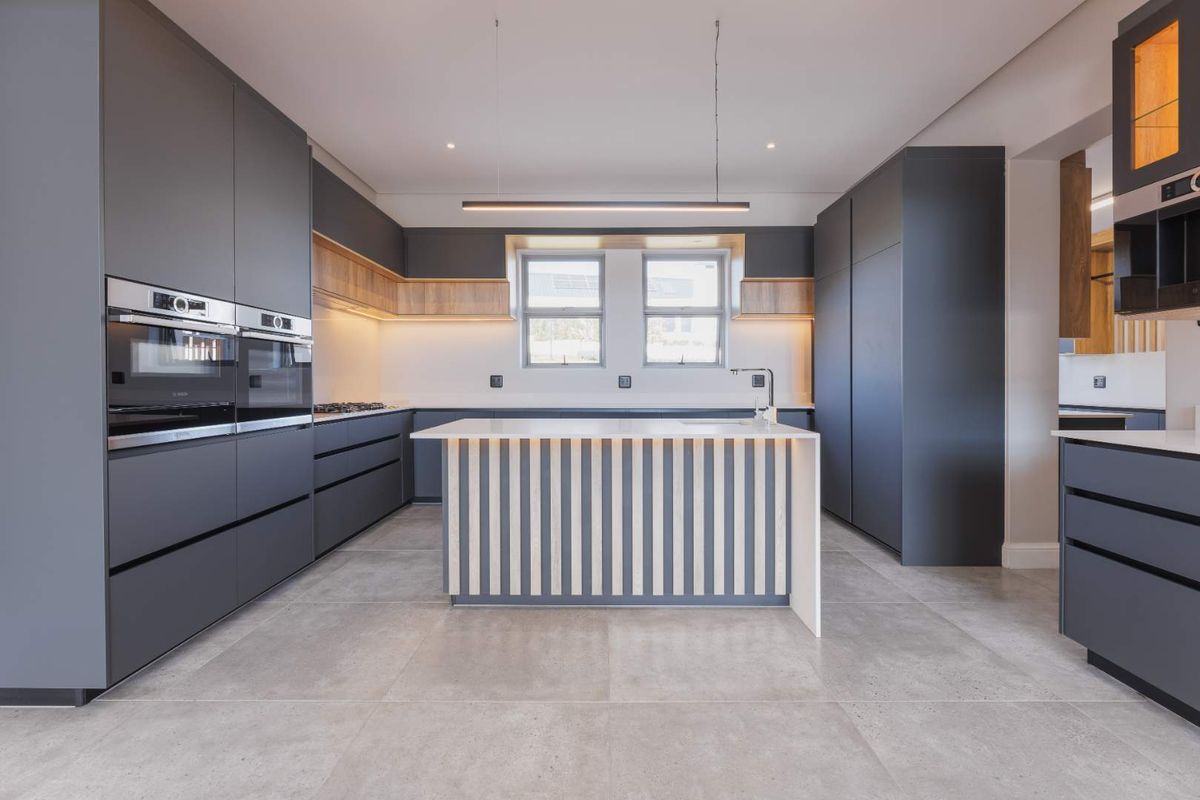Modern kitchen featuring dark matte cabinetry, integrated double ovens, a central island with timber slat detailing, and large format floor tiles.