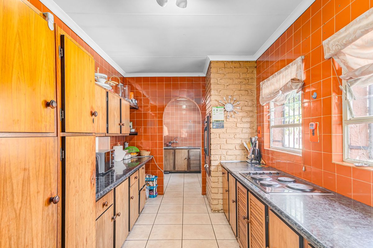 A dated kitchen featuring extensive orange wall tiling, wooden cabinetry, an exposed brick wall, and a separate scullery area with a sink.