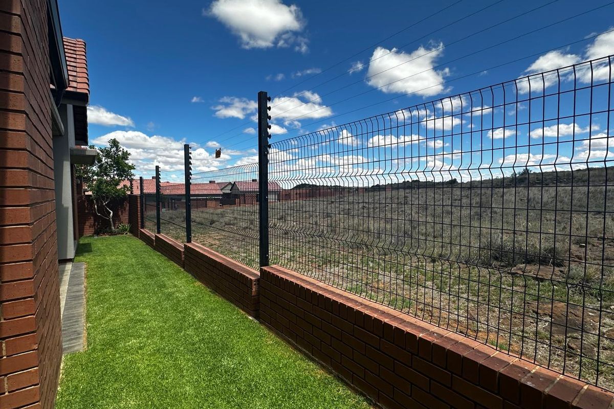 A well-maintained side garden area featuring a brick boundary wall topped with secure wire mesh fencing and integrated electric security wiring.