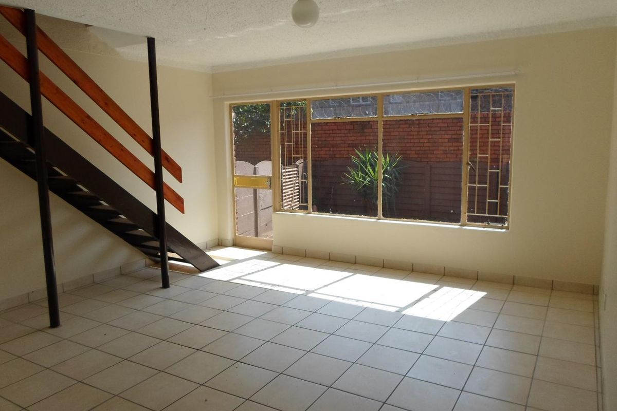 An empty, open-plan living area featuring tiled flooring and a staircase, with direct access to an outdoor space through a security-fitted door.