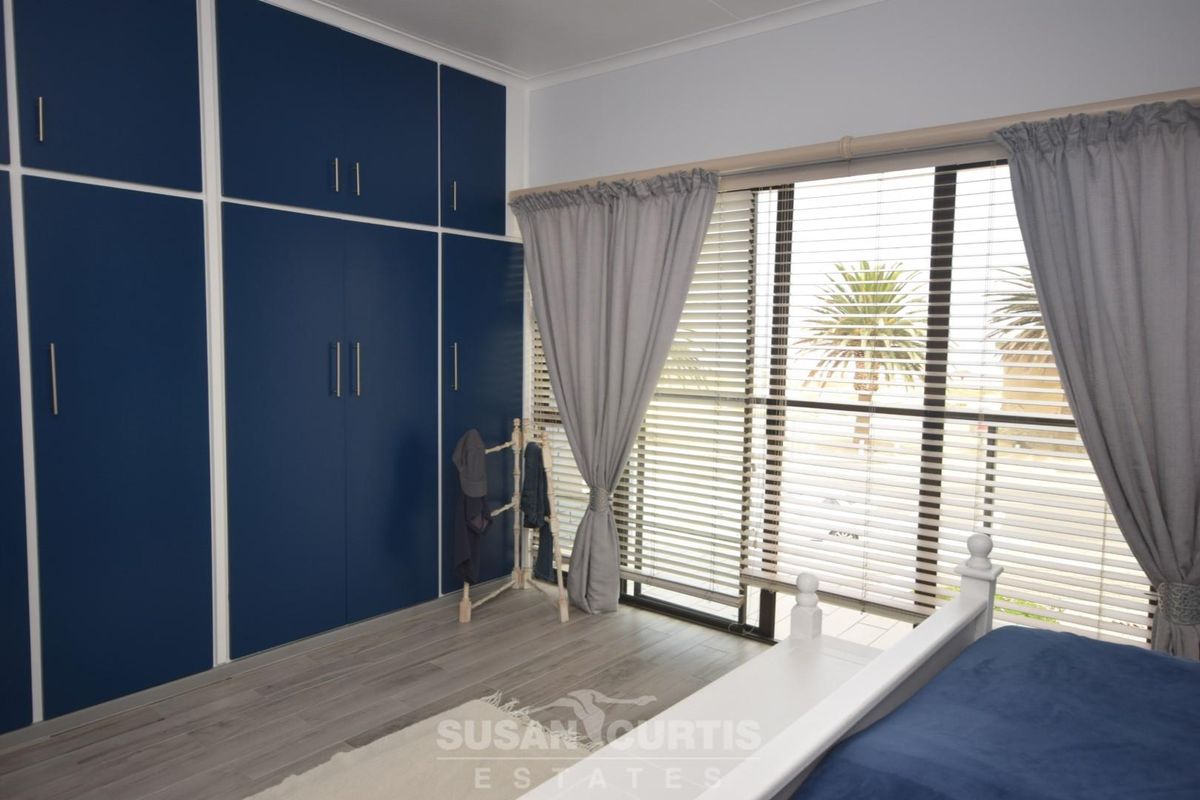 A bright bedroom featuring extensive floor-to-ceiling built-in cabinetry with a modern blue finish, light wood-look flooring, and large windows with integrated blinds.