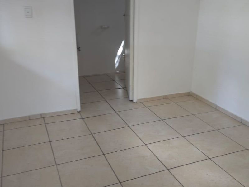 An empty bedroom featuring neutral tiled flooring and a window with a fitted venetian blind, leading into an adjacent room.