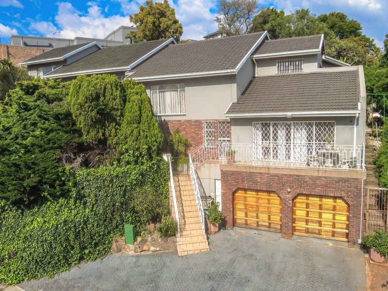 A multi-level residential property featuring a double garage, paved driveway, and an elevated balcony with security gates, set against a backdrop of mature trees.