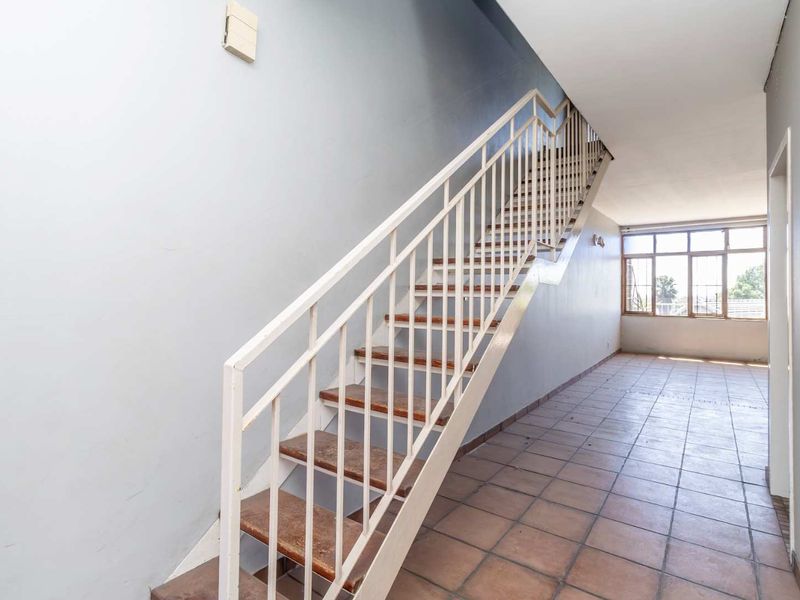 A spacious, empty hallway featuring a central staircase with a metal balustrade, tiled flooring, and natural light from a large window.