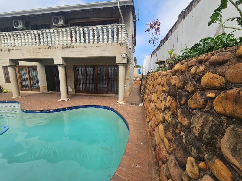 Rear exterior view of the townhouse featuring a swimming pool, brick paved patio, stone retaining wall, and an upper-level balcony with balustrade.