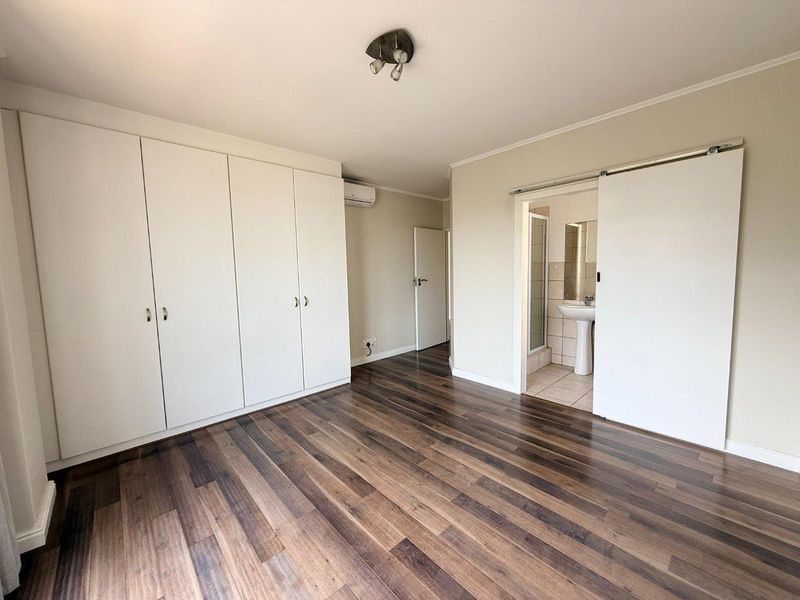 Spacious bedroom featuring laminate flooring, extensive built-in cupboards, an air conditioning unit, and an en-suite bathroom accessed via a sliding door.