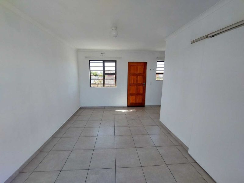 A bright, empty living area featuring neutral tiled flooring, a wooden entrance door, and security bars on the windows.