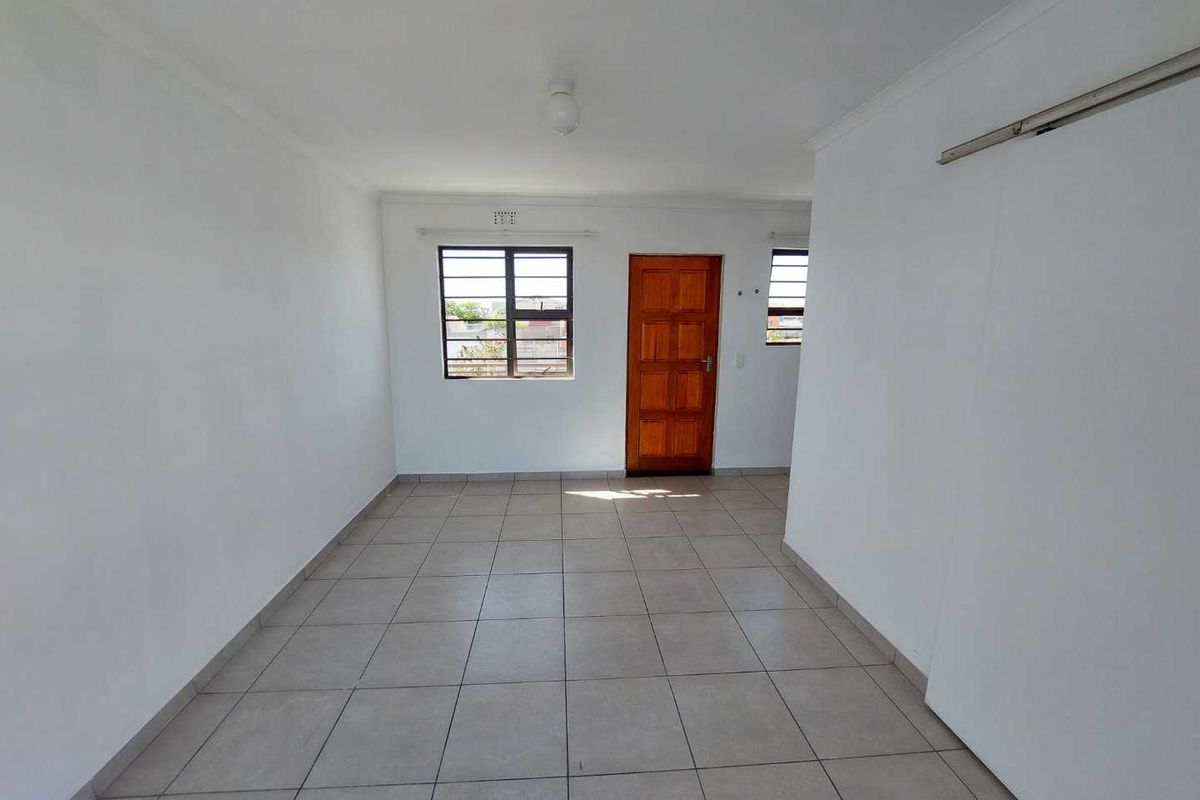 A bright, empty living area featuring neutral tiled flooring, a wooden entrance door, and security bars on the windows.