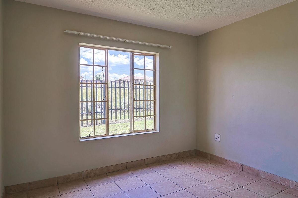 A bright, empty bedroom featuring neutral wall tones, tiled flooring, and a large window with security bars overlooking the exterior.