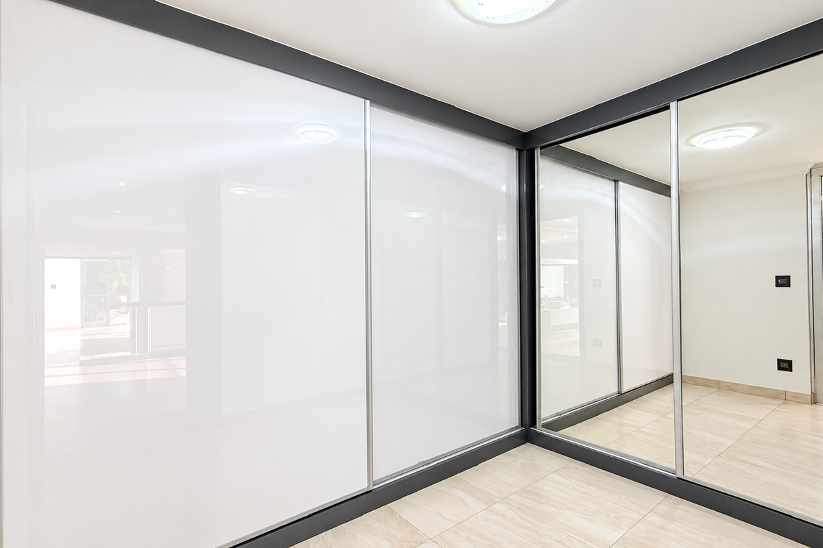 A modern, empty bedroom featuring large built-in sliding wardrobes with mirrored panels, sleek dark frames, and neutral tiled flooring.