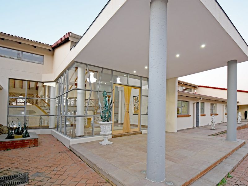 A modern architectural front entrance featuring a large covered portico, extensive glass facade, and paved driveway leading to double wooden doors.