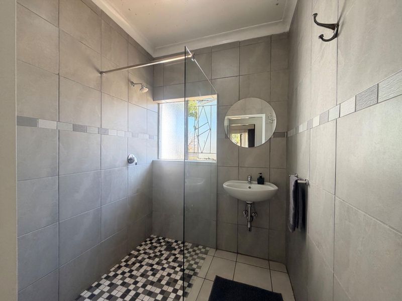 A modern bathroom featuring a walk-in shower with a glass screen, wall-mounted basin, and neutral grey wall tiling with a mosaic floor accent.