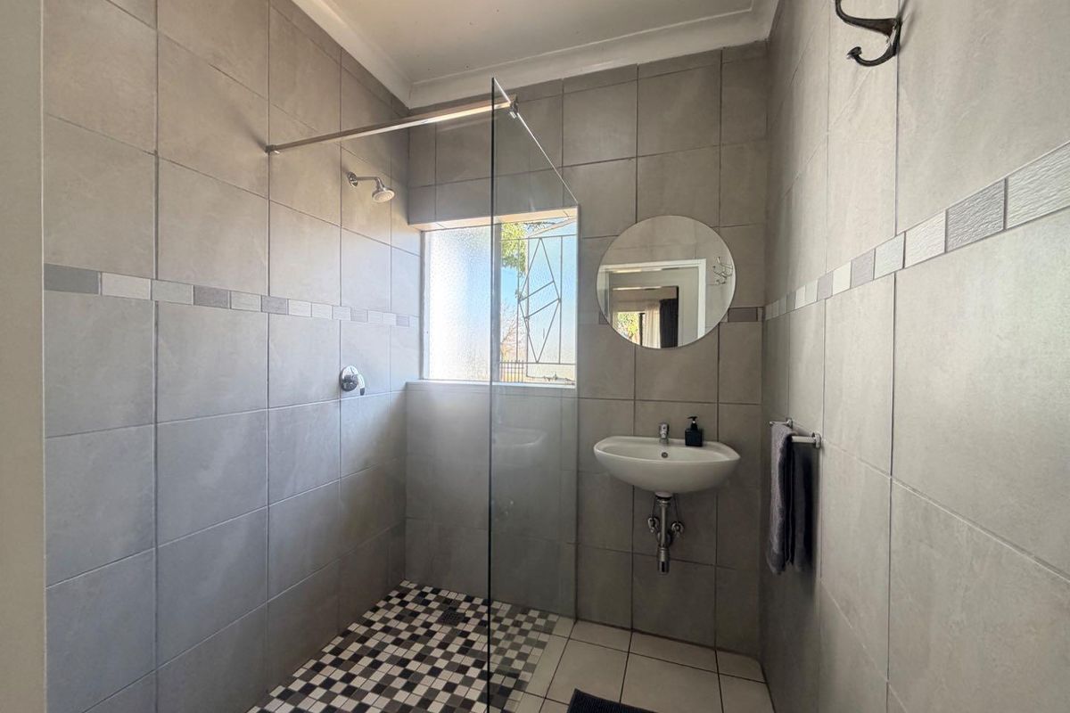 A modern bathroom featuring a walk-in shower with a glass screen, wall-mounted basin, and neutral grey wall tiling with a mosaic floor accent.